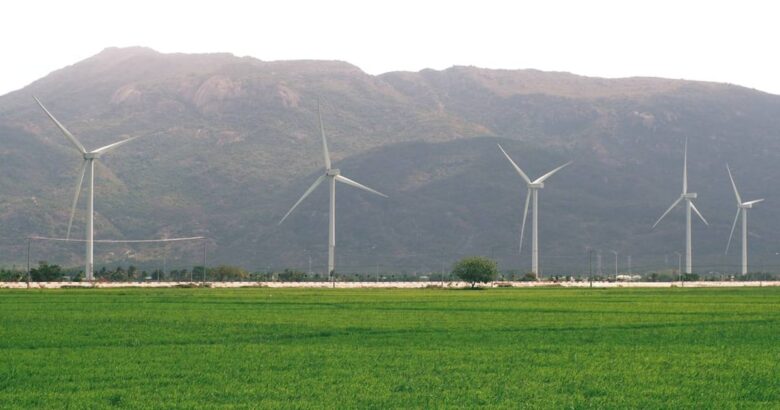 Five wind turbines stand in a green field before mountains.